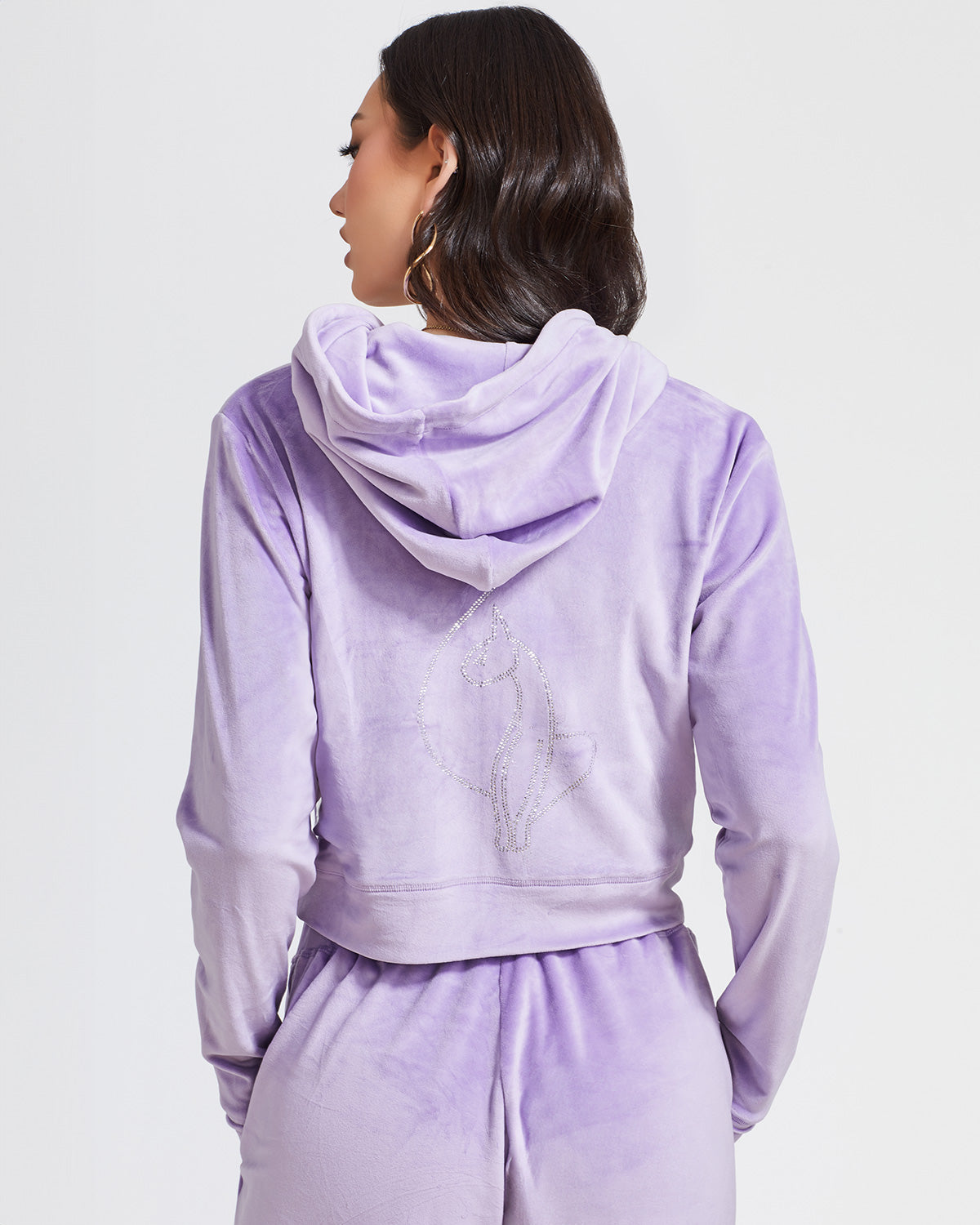 Back view image of standard sized model shown wearing lavender velour zip up hoodie with matching wide leg sweatpants. Zippered sweatshirt features rhinestone Baby Phat script logo on upper chest and oversized rhinestone cat logo on the back. Sweatshirt features silver cat logo zipper pull.