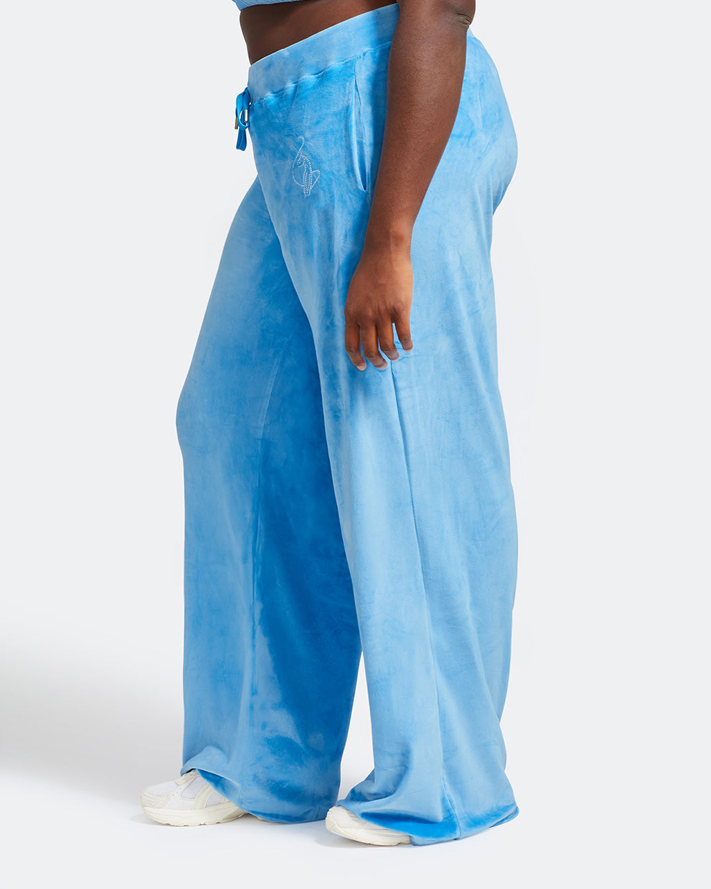 Side view image of plus sized model shown wearing blue velour sweatpants. Pants feature a wide leg, open bottom fit with a rhinestone Baby Phat cat logo on the wearer's left upper thigh. Pants feature pockets and an elastic waistband with a drawstring tie.