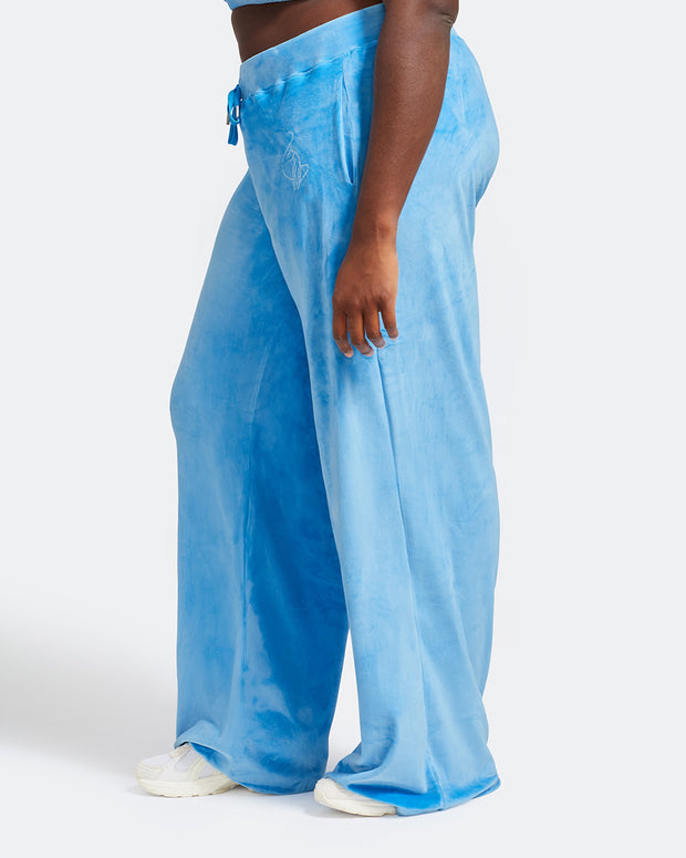 Side view image of plus sized model shown wearing blue velour sweatpants. Pants feature a wide leg, open bottom fit with a rhinestone Baby Phat cat logo on the wearer's left upper thigh. Pants feature pockets and an elastic waistband with a drawstring tie.