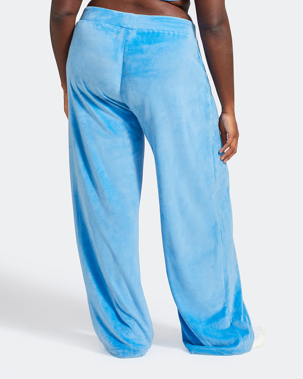 Back view image of plus sized model shown wearing blue velour sweatpants. Pants feature a wide leg, open bottom fit with a rhinestone Baby Phat cat logo on the wearer's left upper thigh. Pants feature pockets and an elastic waistband with a drawstring tie.