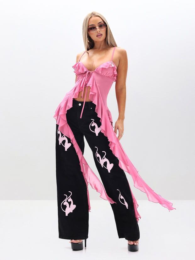 A woman in sunglasses wears the Cami Mesh Waterfall Ruffle Top, a pink piece with long, flowing tails, paired with black pants featuring pink abstract designs, standing against a plain light background.
