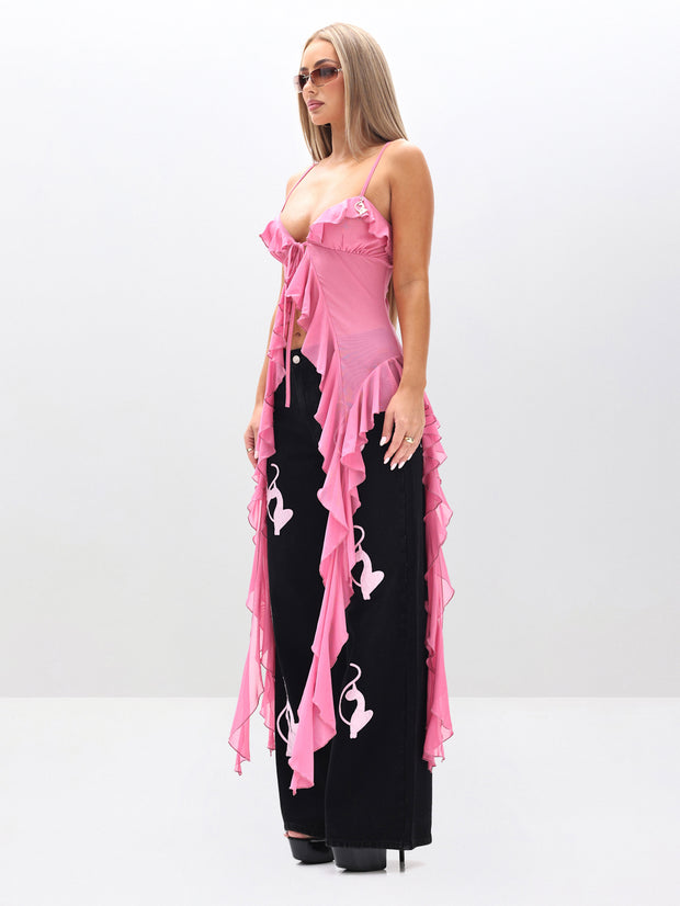 A woman stands against a plain white background wearing sunglasses, the Cami Mesh Waterfall Ruffle Top in pink, and black wide-leg pants featuring white abstract face designs.