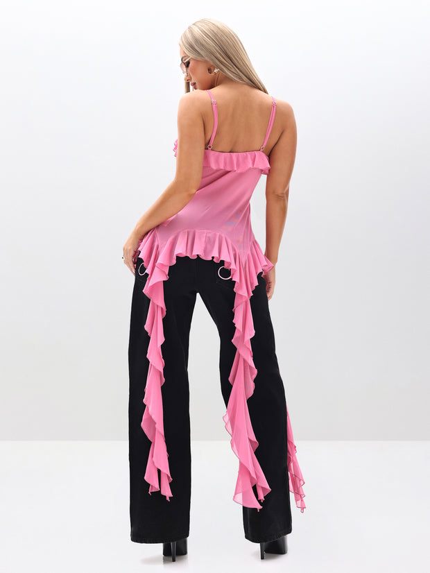 A woman stands facing away in the Cami Mesh Waterfall Ruffle Top, featuring pink ruffles with long side panels, paired with black pants and black platform heels against a plain light background.