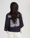 Back view image shows standard sized model wearing black oversized jersey with black rhinestones all over. Jersey features grey 99 logo large at center back with grey rhinestones. Jersey features large grey Baby Phat varsity logo across upper back.