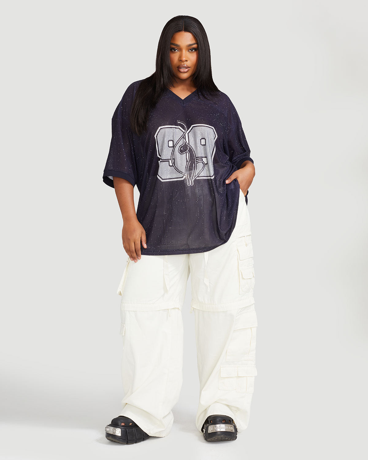 Full body image shows plus sized model wearing black oversized jersey with black rhinestones all over. Jersey features grey 99 cat logo large at center front with grey all over rhinestones. Jersey features v-neckline. Model is also wearing off white cargo pants.