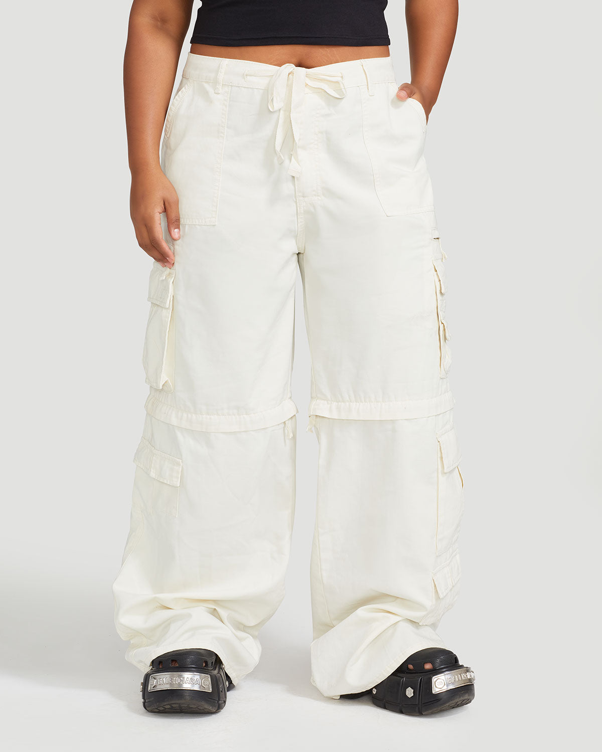 Front view image of plus sized model’s legs wearing off white cargo pant with tied waist and belt loops. Pants feature exaggerated pocket detailing down both legs and hidden zippers below the knees. 