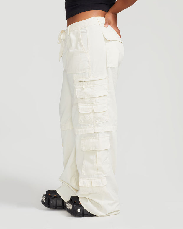 Side view image of plus sized model’s legs wearing off white cargo pant with tied waist and belt loops. Pants feature exaggerated pocket detailing down both legs and large pockets on both sides of rear. Hidden zippers below the knees. 