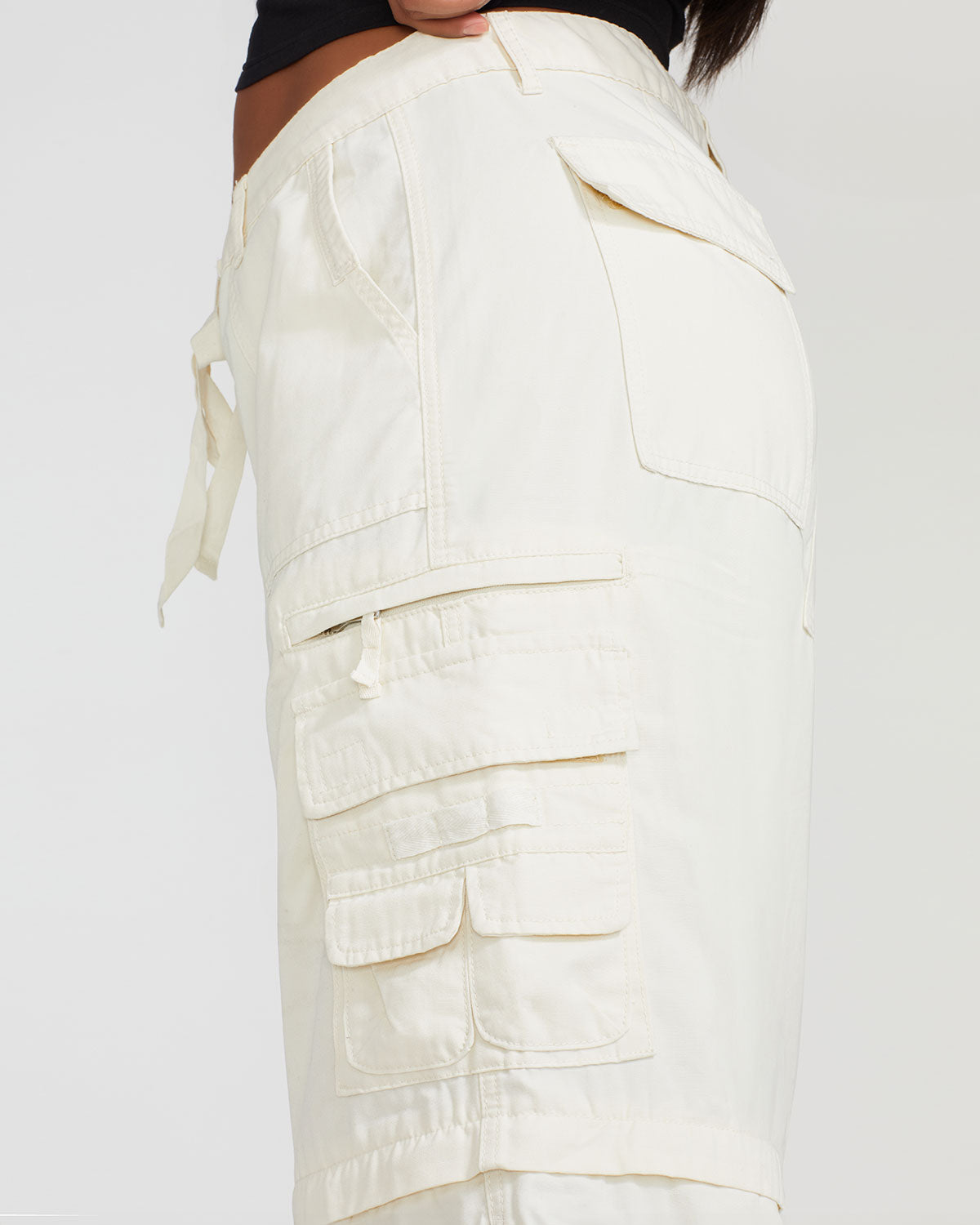 Side view image of plus sized model’s legs wearing off white cargo pant with tied waist and belt loops. Pants feature exaggerated pocket detailing down both legs and large pockets on both sides of rear. 