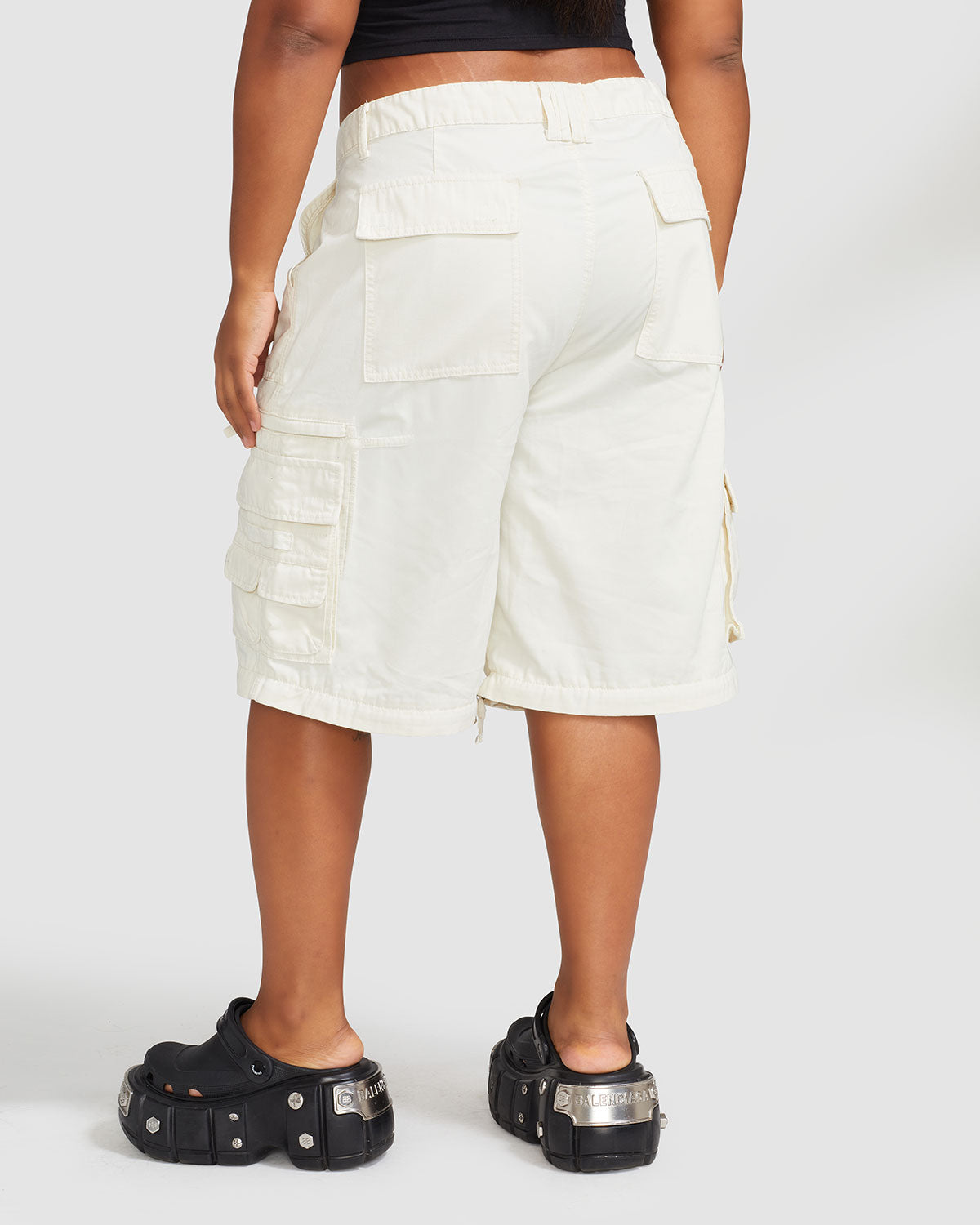 Back view image of plus sized model’s legs wearing off white cargo short with visible belt loops. Shorts feature exaggerated pocket detailing down both legs and 2 oversized pockets at rear.