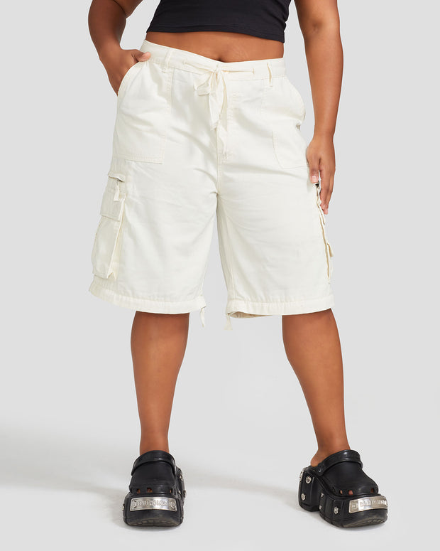 Front view image of plus sized model’s legs wearing off white cargo short with visible belt loops and tied waist. Shorts feature exaggerated pocket detailing down both legs.