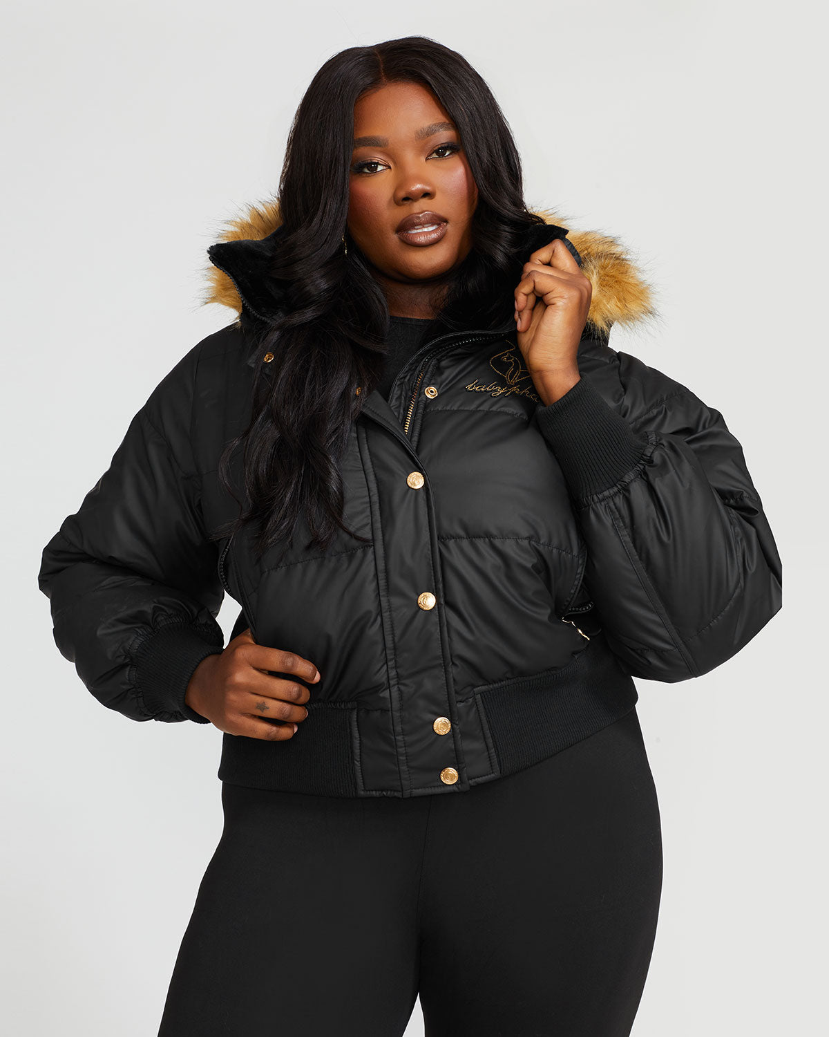 Plus sized model shown wearing black puffer jacket. Coat features natural faux fur trimmed detachable hood and Baby Phat script logo on front chest and oversized cat logo on the back. Jacket also features gold cat logo zipper pulls on the front pockets and center front.