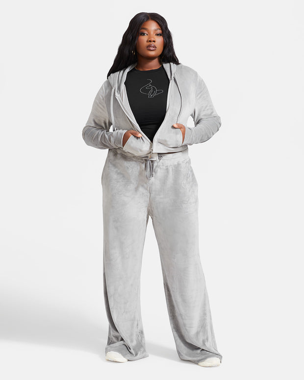 Plus sized model shown wearing grey velour sweatpants. Pants feature a wide leg, open bottom fit with a rhinestone Baby Phat cat logo on the wearer's left upper thigh. Pants feature pockets and an elastic waistband with a drawstring tie. Model is also wearing a black t-shirt with a rhinestone cat logo and a matching grey velour zippered sweatshirt.