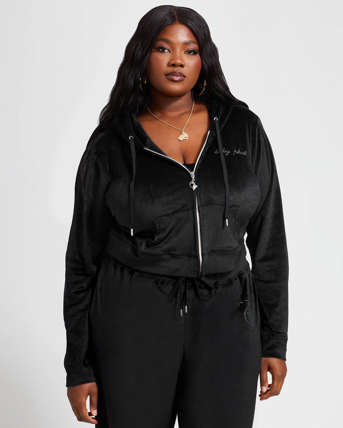 Plus sized model shown wearing black velour zip up hoodie with matching wide leg sweatpants. Zippered sweatshirt features rhinestone Baby Phat script logo on upper chest and oversized rhinestone cat logo on the back. Sweatshirt features silver cat logo zipper pull.