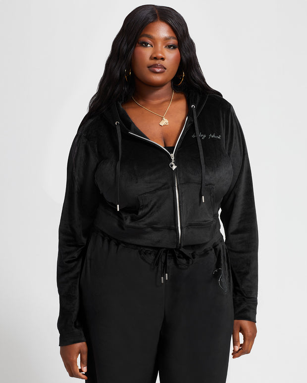 Plus sized model shown wearing black velour zip up hoodie with matching wide leg sweatpants. Zippered sweatshirt features rhinestone Baby Phat script logo on upper chest and oversized rhinestone cat logo on the back. Sweatshirt features silver cat logo zipper pull.