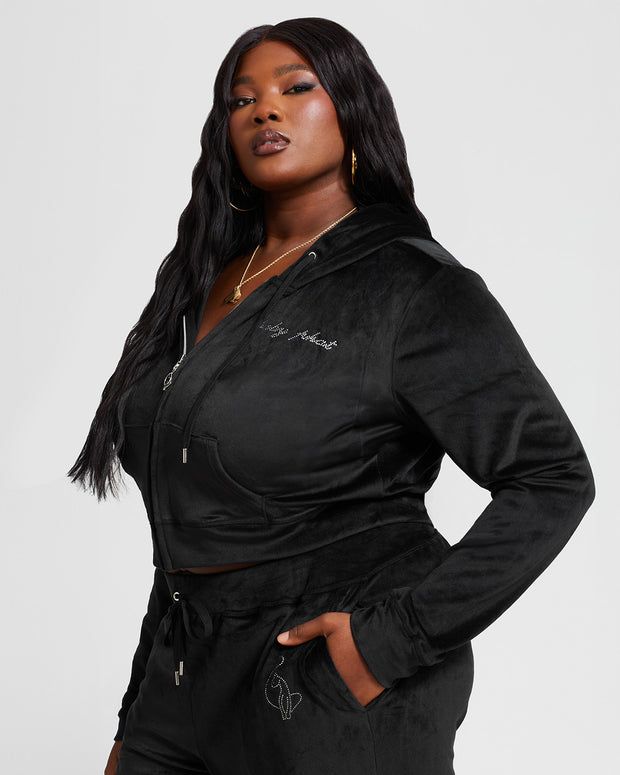 Plus sized model shown wearing black velour zip up hoodie with matching wide leg sweatpants. Zippered sweatshirt features rhinestone Baby Phat script logo on upper chest and oversized rhinestone cat logo on the back. Sweatshirt features silver cat logo zipper pull.