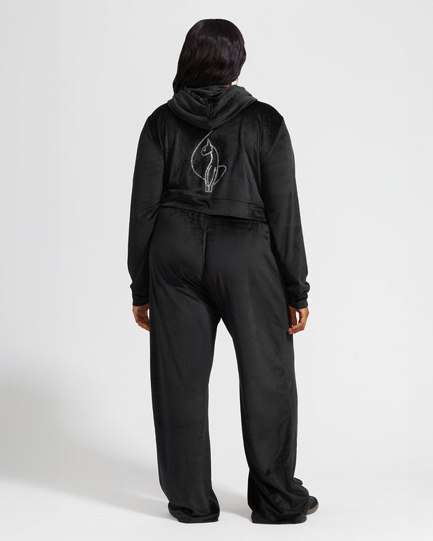 Full length back view image of plus sized model shown wearing black velour zip up hoodie with matching wide leg sweatpants. Zippered sweatshirt features rhinestone Baby Phat script logo on upper chest and oversized rhinestone cat logo on the back. Sweatshirt features silver cat logo zipper pull.