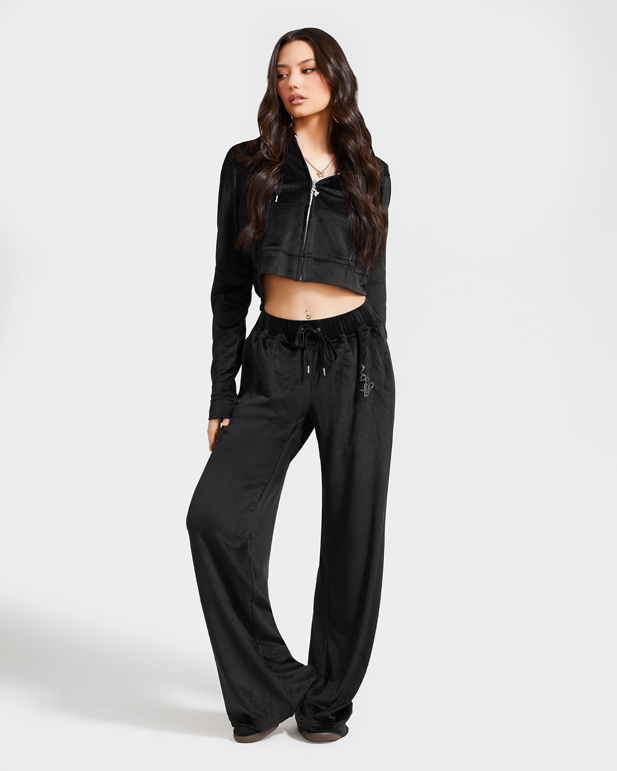 Full length image of standard sized model shown wearing black velour zip up hoodie with matching wide leg sweatpants. Zippered sweatshirt features rhinestone Baby Phat script logo on upper chest and oversized rhinestone cat logo on the back. Sweatshirt features silver cat logo zipper pull.