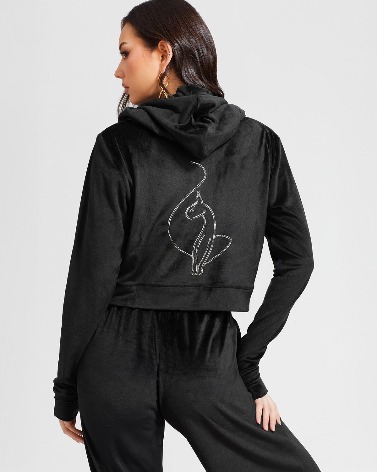 Back view image of standard sized model shown wearing black velour zip up hoodie with matching wide leg sweatpants. Zippered sweatshirt features rhinestone Baby Phat script logo on upper chest and oversized rhinestone cat logo on the back. Sweatshirt features silver cat logo zipper pull.