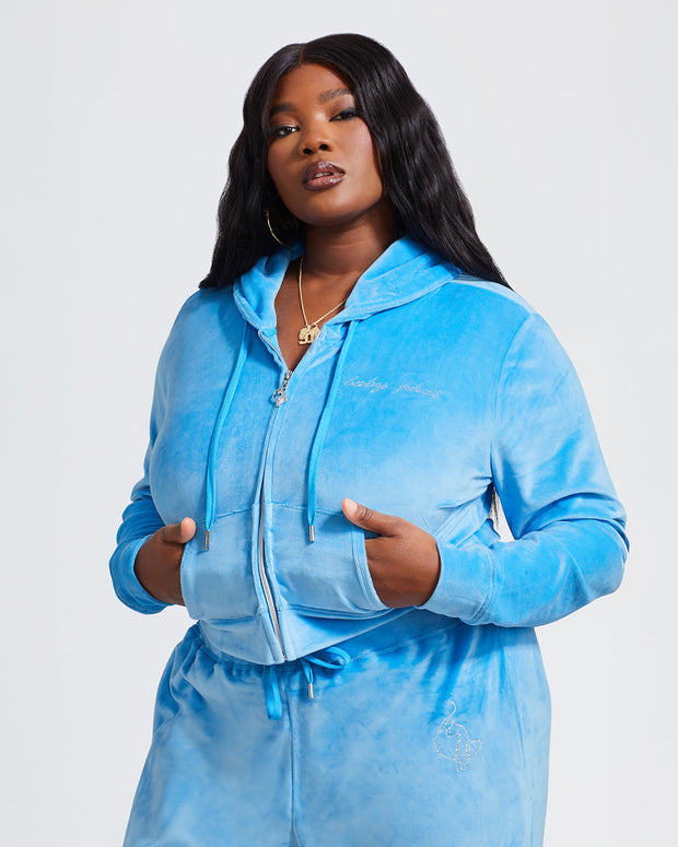Plus sized model shown wearing blue velour zip up hoodie with matching wide leg sweatpants. Zippered sweatshirt features rhinestone Baby Phat script logo on upper chest and oversized rhinestone cat logo on the back. Sweatshirt features silver cat logo zipper pull.