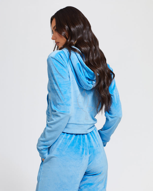 Back view image of standard sized model shown wearing blue velour zip up hoodie with matching wide leg sweatpants. Zippered sweatshirt features rhinestone Baby Phat script logo on upper chest and oversized rhinestone cat logo on the back. Sweatshirt features silver cat logo zipper pull.