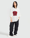 Full body image shows standard sized model wearing off white oversized jersey with clear rhinestones all over. Jersey features burgundy 99 cat logo large at center front with burgundy all over rhinestones. Jersey features v-neckline. Model is also wearing black cargo pants.
