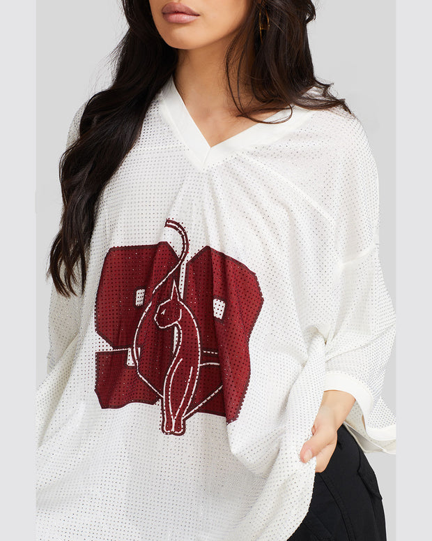 Detail image shows standard sized model wearing off white oversized jersey with clear rhinestones all over. Jersey features burgundy 99 cat logo large at center front with burgundy all over rhinestones. Jersey features v-neckline. Model is also wearing black cargo pants.
