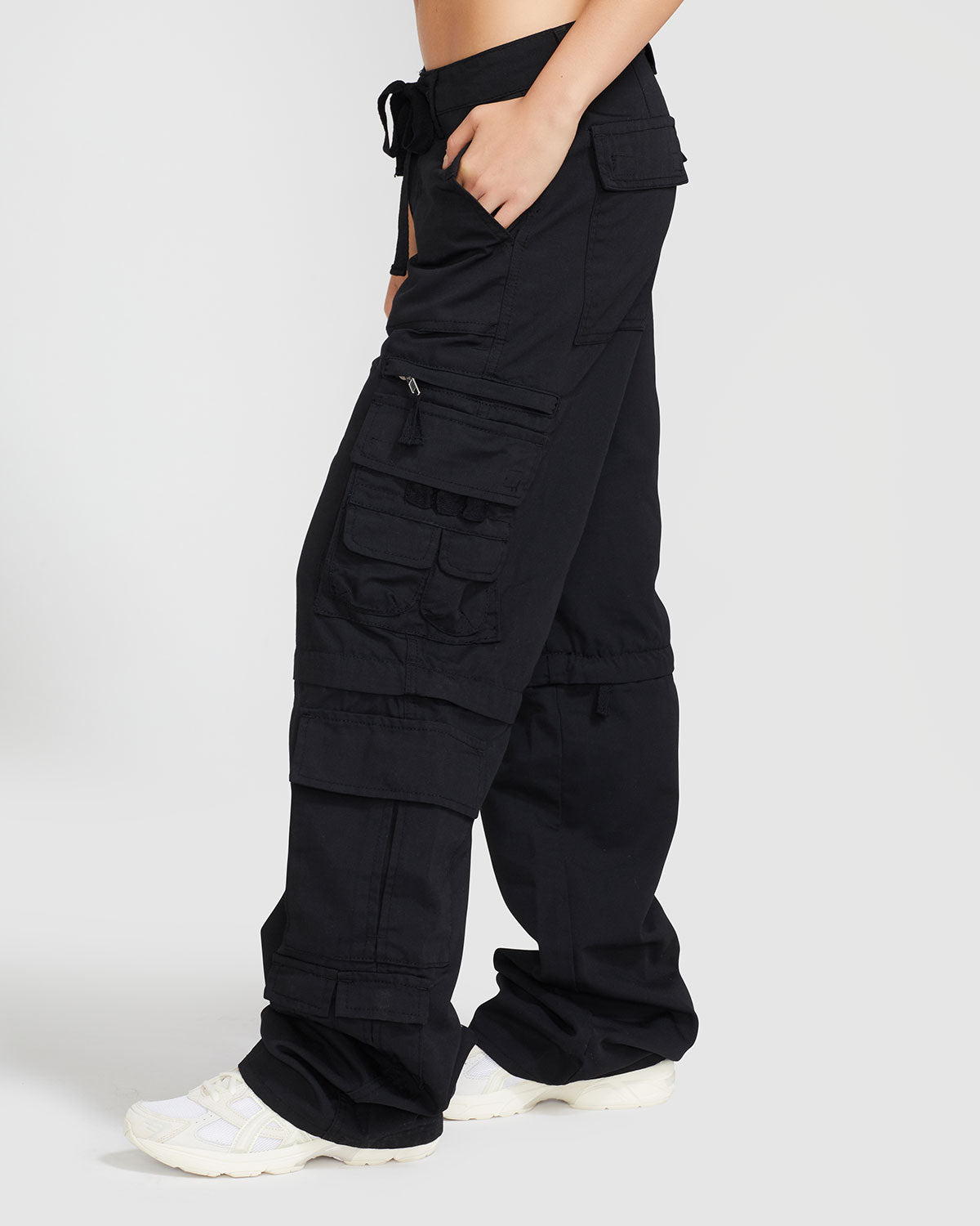 Side view image of standard sized model’s legs wearing black cargo pant with tied waist and belt loops. Pants feature exaggerated pocket detailing down both legs and hidden zippers below the knees. 