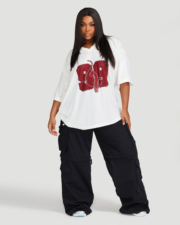 Full body image shows plus sized model wearing off white oversized jersey with clear rhinestones all over. Jersey features burgundy 99 cat logo large at center front with burgundy all over rhinestones. Jersey features v-neckline. Model is also wearing black cargo pants.