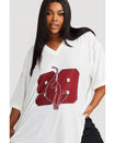 Plus sized model shown wearing off white oversized jersey with clear rhinestones all over. Jersey features burgundy 99 cat logo large at center front with burgundy all over rhinestones.  Jersey features v-neckline. Model is also wearing black cargo pants.