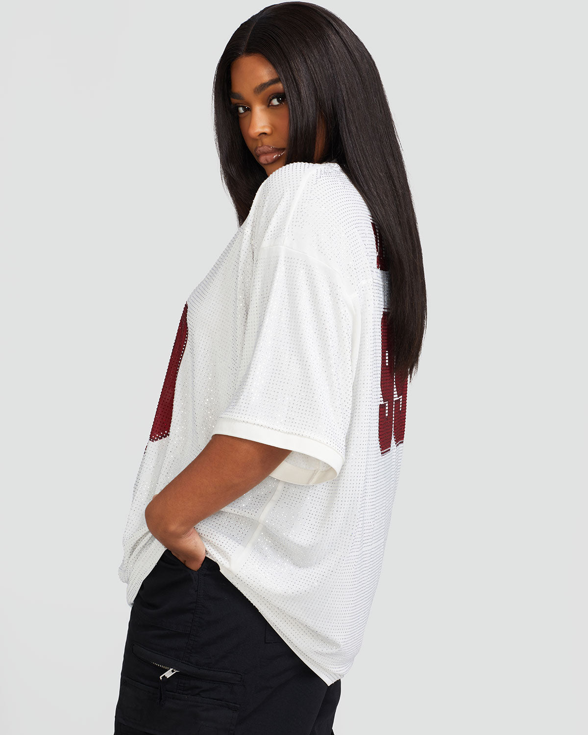 Side view image shows plus sized model wearing off white oversized jersey with clear rhinestones all over. Jersey features burgundy 99 cat logo large at center front with burgundy all over rhinestones. Jersey features v-neckline. Model is also wearing black cargo pants.
