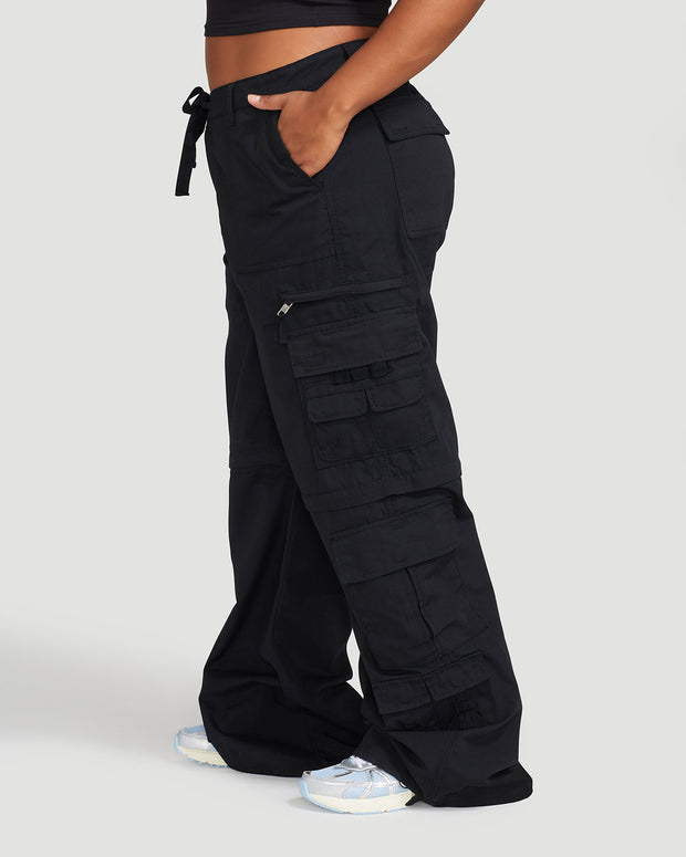 Side view image of plus sized model’s legs wearing black cargo pant with tied waist and belt loops. Pants feature exaggerated pocket detailing down both legs and hidden zippers below the knees. 