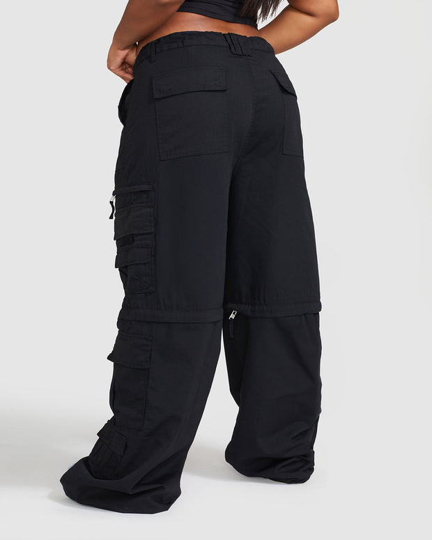 Back view image of plus sized model’s legs wearing black cargo pant with visible belt loops. Pants feature exaggerated pocket detailing down both legs and large pockets on both sides of rear. Hidden zippers below the knees. 
