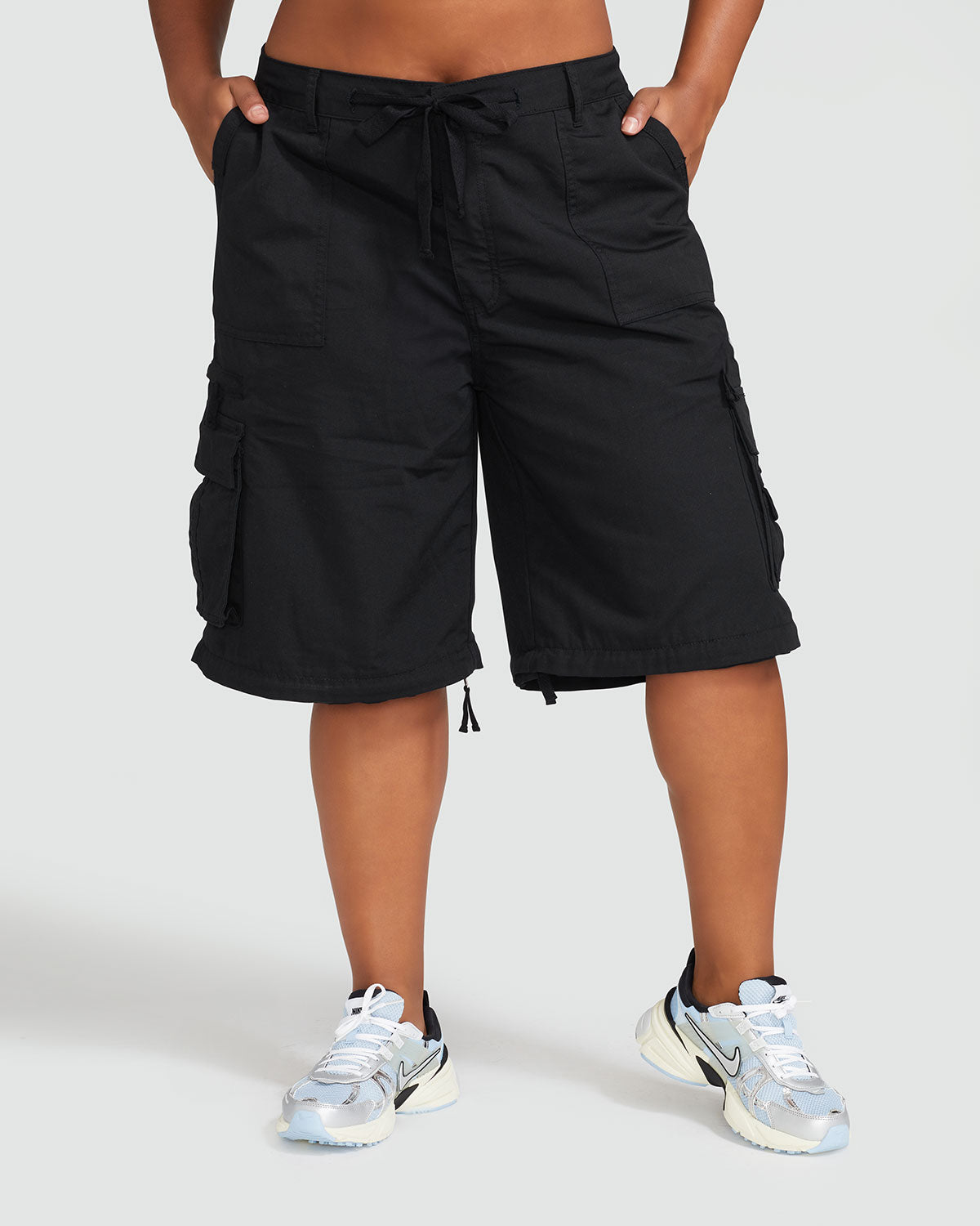 Front view image of standard sized model’s legs wearing black cargo short with visible belt loops and tied waist. Shorts feature exaggerated pocket detailing down both legs. 