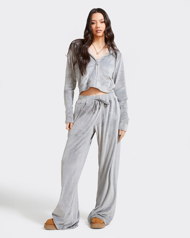 Full length image of standard sized model shown wearing grey velour sweatpants. Pants feature a wide leg, open bottom fit with a rhinestone Baby Phat cat logo on the wearer's left upper thigh. Pants feature pockets and an elastic waistband with a drawstring tie. Model is shown wearing matching grey velour zippered sweatshirt.