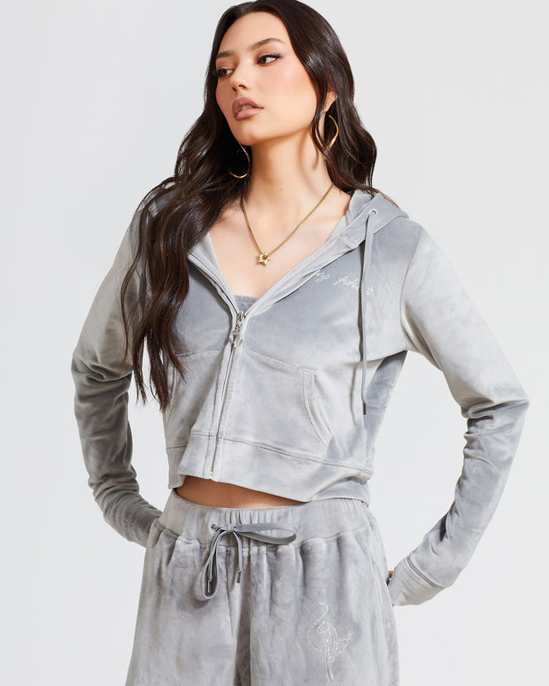 Standard sized model shown wearing grey velour zip up hoodie with matching wide leg sweatpants. Zippered sweatshirt features rhinestone Baby Phat script logo on upper chest and oversized rhinestone cat logo on the back. Sweatshirt features silver cat logo zipper pull.