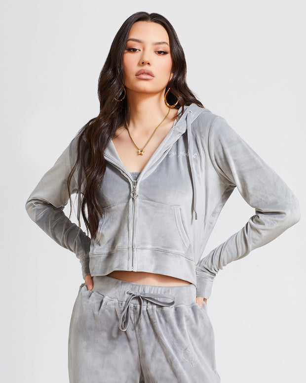 Standard sized model shown wearing grey velour zip up hoodie with matching wide leg sweatpants. Zippered sweatshirt features rhinestone Baby Phat script logo on upper chest and oversized rhinestone cat logo on the back. Sweatshirt features silver cat logo zipper pull.