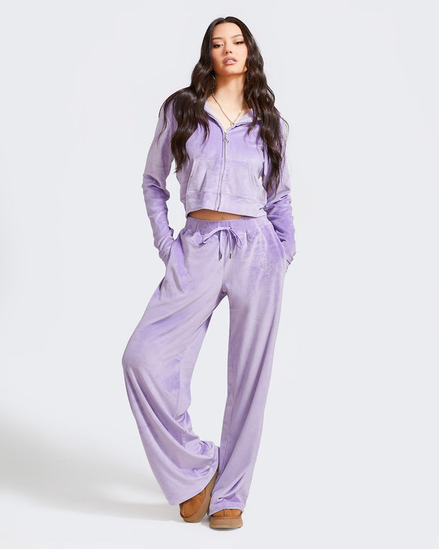 Full length image of standard sized model shown wearing light purple velour sweatpants. Pants feature a wide leg, open bottom fit with a rhinestone Baby Phat cat logo on the wearer's left upper thigh. Pants feature pockets and an elastic waistband with a drawstring tie. Model is also wearing a matching zippered sweatshirt.