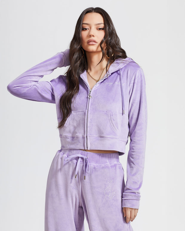 Standard sized model shown wearing lavender velour zip up hoodie with matching wide leg sweatpants. Zippered sweatshirt features rhinestone Baby Phat script logo on upper chest and oversized rhinestone cat logo on the back. Sweatshirt features silver cat logo zipper pull.