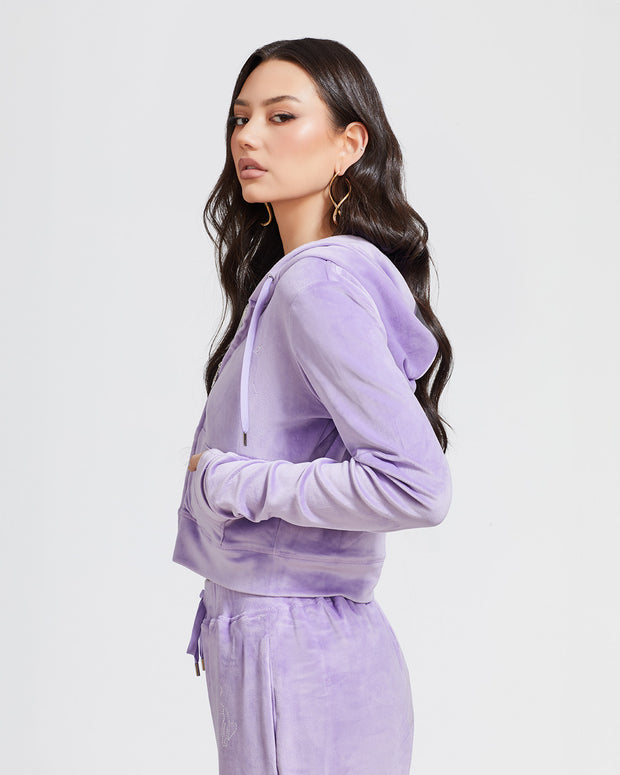 Side view image of standard sized model shown wearing lavender velour zip up hoodie with matching wide leg sweatpants. Zippered sweatshirt features rhinestone Baby Phat script logo on upper chest and oversized rhinestone cat logo on the back. Sweatshirt features silver cat logo zipper pull.