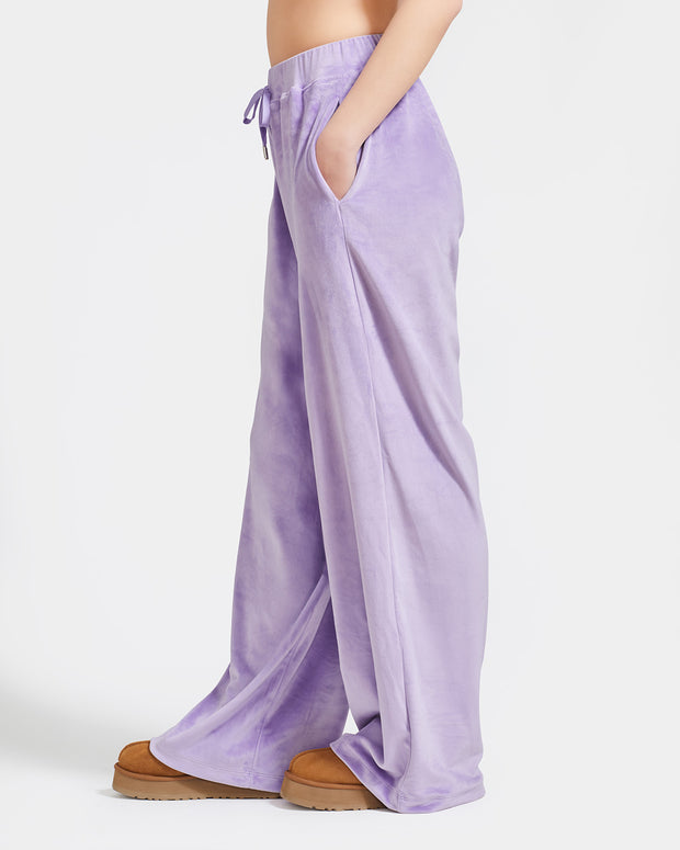 Side view image of standard sized model shown wearing light purple velour sweatpants. Pants feature a wide leg, open bottom fit with a rhinestone Baby Phat cat logo on the wearer's left upper thigh. Pants feature pockets and an elastic waistband with a drawstring tie.