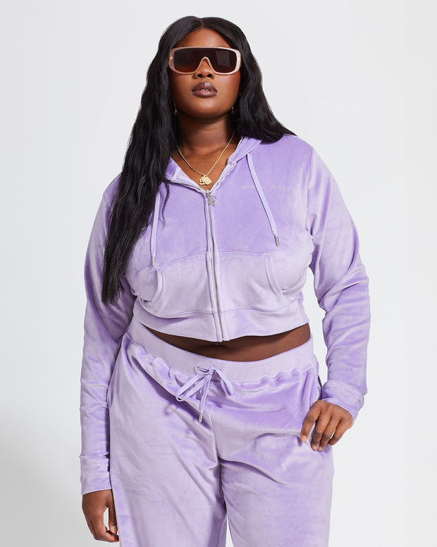 Plus sized model shown wearing lavender velour zip up hoodie with matching wide leg sweatpants. Zippered sweatshirt features rhinestone Baby Phat script logo on upper chest and oversized rhinestone cat logo on the back. Sweatshirt features silver cat logo zipper pull.