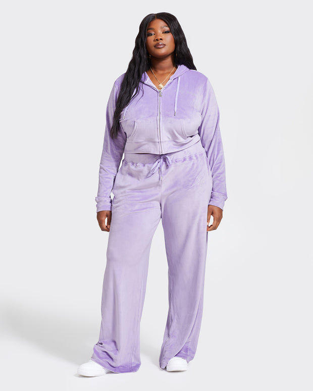 Full body image of plus sized model shown wearing lavender velour zip up hoodie with matching wide leg sweatpants. Zippered sweatshirt features rhinestone Baby Phat script logo on upper chest and oversized rhinestone cat logo on the back. Sweatshirt features silver cat logo zipper pull.