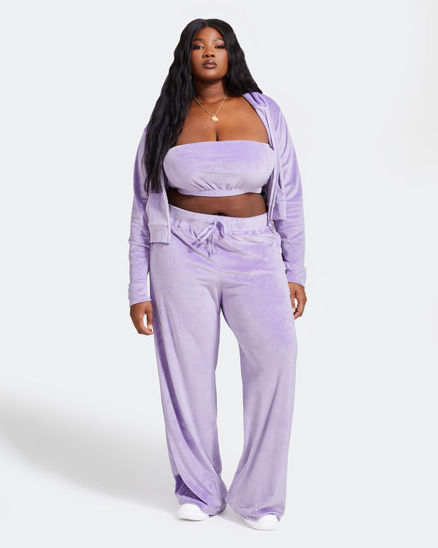 Full length image of plus sized model shown wearing light purple velour sweatpants. Pants feature a wide leg, open bottom fit with a rhinestone Baby Phat cat logo on the wearer's left upper thigh. Pants feature pockets and an elastic waistband with a drawstring tie. Model is also wearing a matching zippered sweatshirt unzipped to show a matching purple velour bandeau.