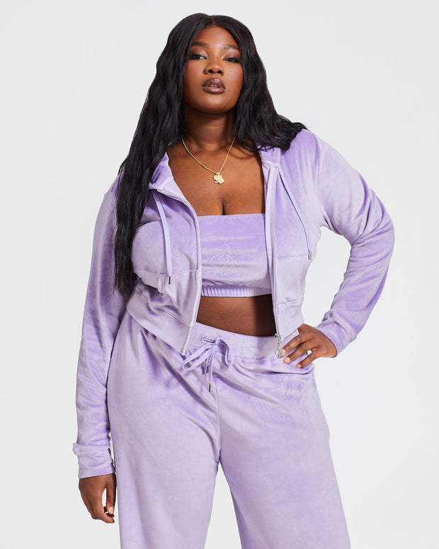 Plus sized model shown wearing lavender velour zip up hoodie open with matching tube top bandeau and wide leg sweatpants. Zippered sweatshirt features rhinestone Baby Phat script logo on upper chest and oversized rhinestone cat logo on the back. Sweatshirt features silver cat logo zipper pull.