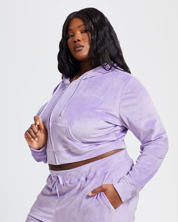 Side view image of plus sized model shown wearing lavender velour zip up hoodie with matching wide leg sweatpants. Zippered sweatshirt features rhinestone Baby Phat script logo on upper chest and oversized rhinestone cat logo on the back. Sweatshirt features silver cat logo zipper pull.
