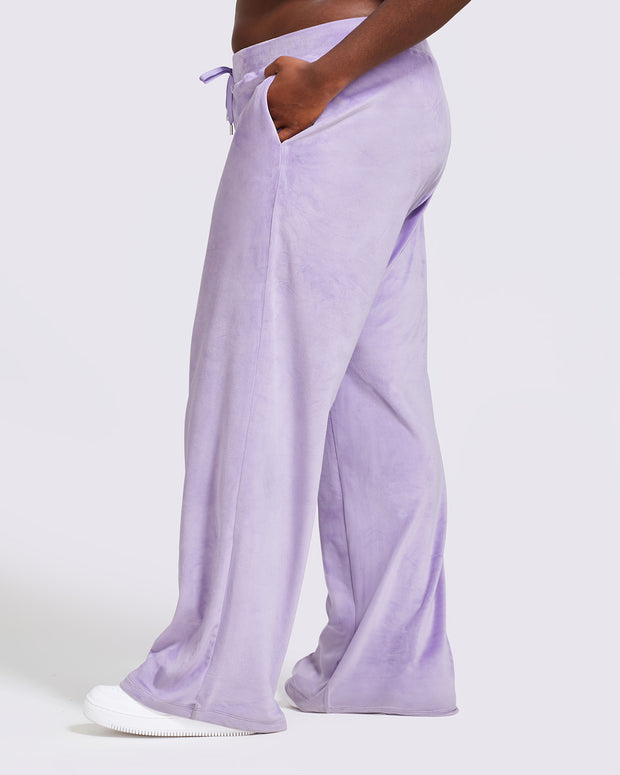 Side view image of plus sized model shown wearing light purple velour sweatpants. Pants feature a wide leg, open bottom fit with a rhinestone Baby Phat cat logo on the wearer's left upper thigh. Pants feature pockets and an elastic waistband with a drawstring tie.