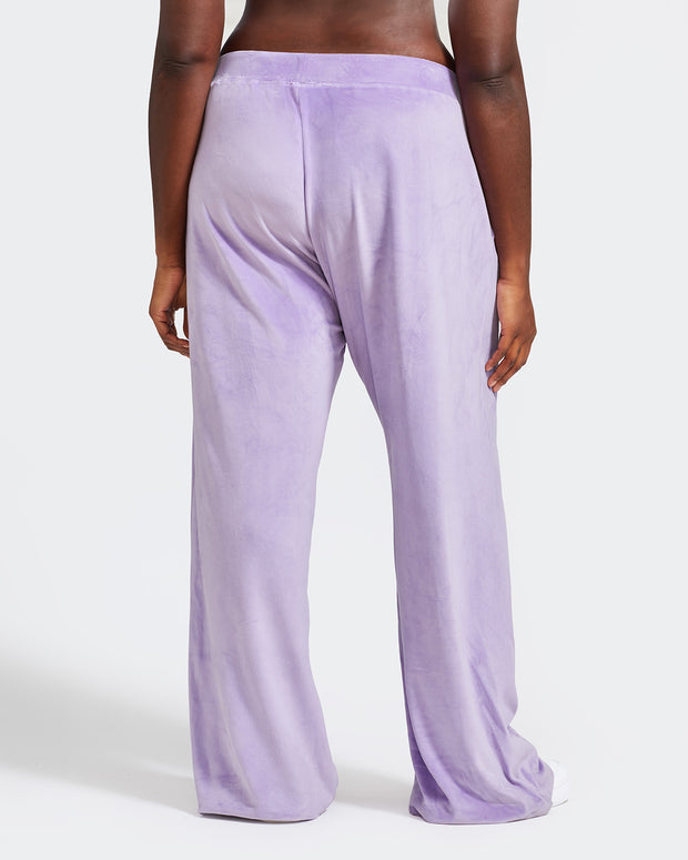 Back view image of plus sized model shown wearing light purple velour sweatpants. Pants feature a wide leg, open bottom fit with a rhinestone Baby Phat cat logo on the wearer's left upper thigh. Pants feature pockets and an elastic waistband with a drawstring tie.