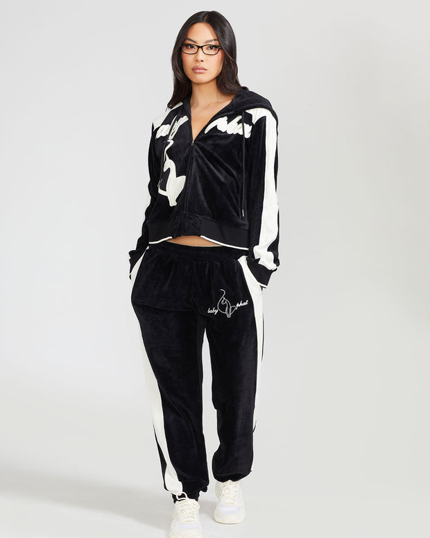 Full body image of standard sized model wearing black velour zip up hoodie with color blocked off white panel down both sleeves. Zippered sweatshirt features off white Baby Phat script logo across upper chest and oversized off white solid cat logo on wearer's right front. Model is also wearing matching black sweatpant with off white color blocking and off white embroidered script and cat logo on wearer's upper left thigh.