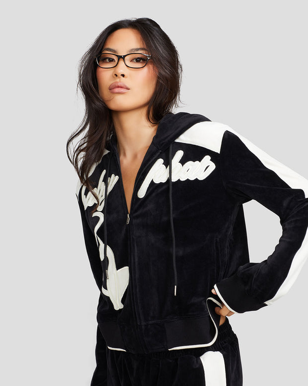 Standard sized model shown wearing black velour zip up hoodie with color blocked off white panel down both sleeves. Zippered sweatshirt features off white Baby Phat script logo across upper chest and oversized solid cat logo on wearer's right front. Sweatshirt features black ribbing at the wrists and hem that has off white tipping.