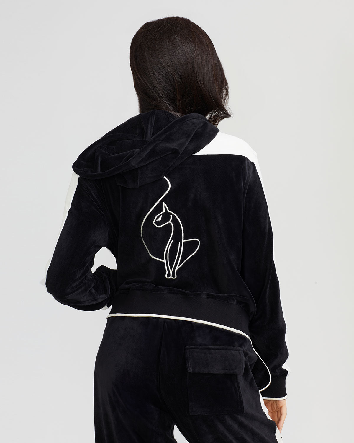 Back view image of standard sized model wearing black velour zip up hoodie with color blocked off white panel down both sleeves. Zippered sweatshirt features matching black ribbing at the wrists and hem with off white tipping. Large embroidered cat logo on center back. Model is also wearing matching black sweatpant with off white color blocking. 